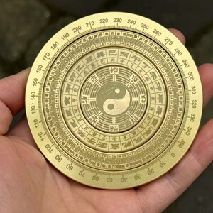 May include: A brass compass with intricate engravings and a Yin Yang symbol at the center. The compass features degree markings and Chinese characters around the perimeter. A traditional tool for navigation and divination.