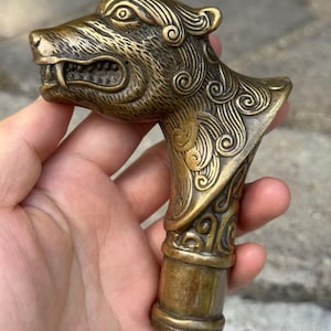 Op de afbeelding: Een metalen wandelstokhandvat in bronskleur in de vorm van een gestileerde wolvenkop. Het ontwerp bevat ingewikkelde wervelpatronen langs de nek en kop, met zichtbare hoektanden en gedetailleerde vachtstructuren. De basis is cilindervormig en glad.