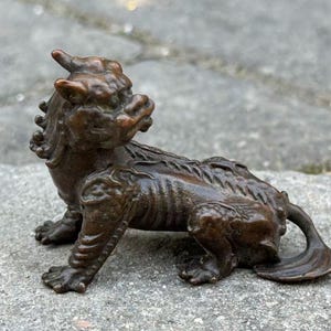 May include: A bronze-colored figurine of a mythical creature, possibly a Foo Dog or similar guardian figure. The sculpture has a detailed, textured surface and is posed in a seated position. The figure has a curled tail and is resting on a stone surface.