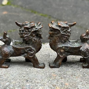 May include: Two bronze-colored, mythical creature figurines. Each figurine has a lion-like body, horns, and a detailed mane. The figurines are positioned facing each other, with intricate details visible on their bodies. They are placed on a textured, gray surface.