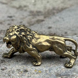 May include: A small, detailed brass lion figurine in a roaring pose. The sculpture has a textured mane and body, with visible claws and a curled tail. The lion is positioned on a rough, gray surface.