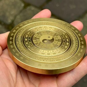 May include: A round, brass-colored compass with intricate markings and a Yin Yang symbol at the center. The compass has degree markings around the edge and is held in a hand.