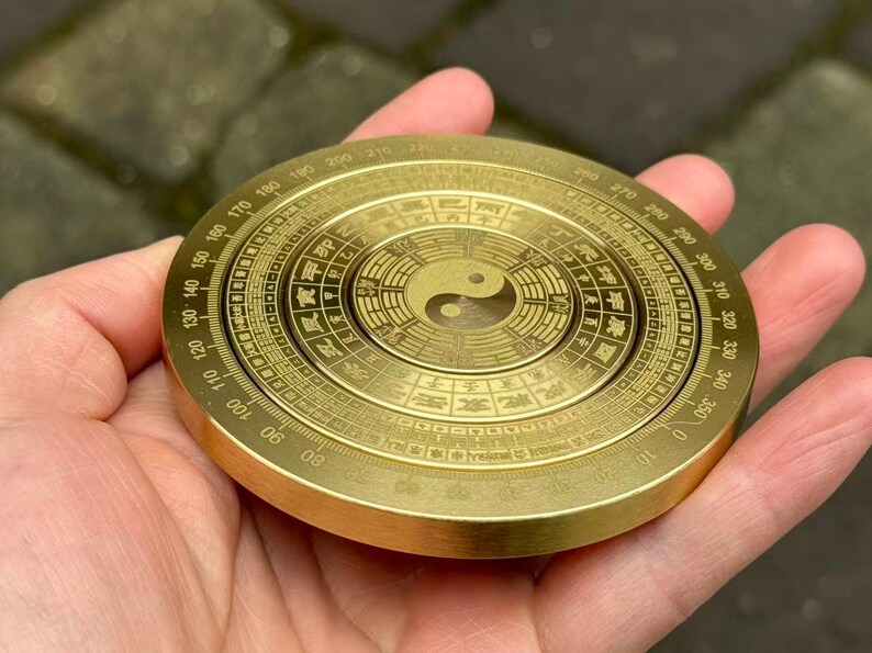 May include: A round, brass-colored compass with intricate markings and a Yin Yang symbol at the center. The compass has degree markings around the edge and is held in a hand.