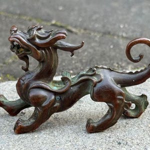 May include: A bronze-colored, mythical creature figurine with a dragon-like appearance. The sculpture features intricate details, including a detailed head, flowing mane, and a curled tail. The figurine is posed in a dynamic stance, with its legs spread.