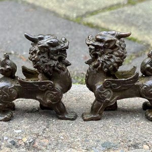 May include: A pair of bronze-colored, mythical creature figurines. Each has a lion-like body with wings, a horned head, and a detailed mane. The figurines are displayed on a stone surface, showcasing their intricate design and antique aesthetic.