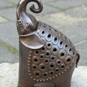 Puede incluir: Una figura de elefante de metal marrón oscuro con la trompa curvada hacia arriba y una zona de silla de montar decorativa cubierta con un patrón de anillos circulares en relieve con centros abiertos. La escultura tiene un acabado liso y una cola.