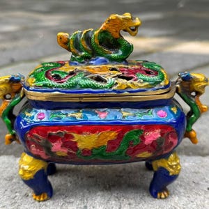 May include: A rectangular metal incense burner featuring vibrant cloisonné enamel work in shades of blue, red, green, and yellow. The lid is topped with a sculpted dragon figure, while the sides are adorned with dragon handles and four legs.