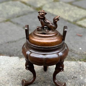 May include: A bronze incense burner with a decorative lid featuring a small, detailed lion figure. The burner has a rounded body supported by three curved legs. The overall design is ornate and antique-inspired.