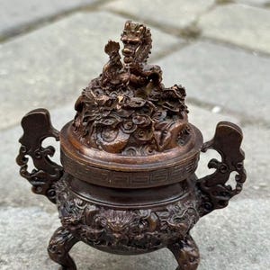 May include: A bronze-colored incense burner with intricate dragon and cloud detailing. The burner features a lid with a dragon sculpture, ornate handles, and three clawed feet. The overall design is detailed and ornate, suggesting a traditional Asian aesthetic.