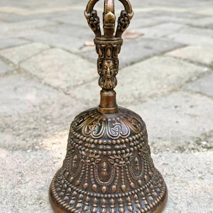 May include: Bronze-colored bell with intricate embossed patterns and a detailed handle. The handle features a decorative top and a carved face. The bell is likely used for spiritual or ceremonial purposes.