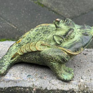 May include: A detailed, green and gold frog figurine with a coin in its mouth. The frog has a textured surface and is adorned with several gold coins along its back. A gold rope-like detail accents the frog's body.