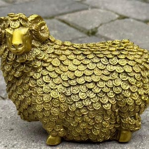 Könnte beinhalten: Eine goldfarbene Widderfigur mit aufwendigen, münzartigen Details, die ihren Körper bedecken. Der Widder hat gebogene Hörner und steht auf vier kleinen Beinen. Die Statue besteht aus glänzendem Metall und steht vor einem Steinhintergrund.