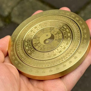 May include: A golden, circular compass with intricate engravings and a central Yin Yang symbol. The compass features degree markings and Chinese characters around the perimeter. The object is held in a hand.