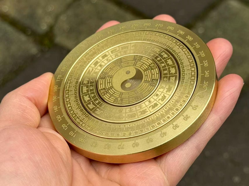 May include: A golden, circular compass with intricate engravings and a central Yin Yang symbol. The compass features degree markings and Chinese characters around the perimeter. The object is held in a hand.