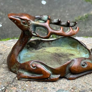 Op de afbeelding: Bronzen hertfiguur met een ingebouwde schaal. Het hert ligt, met het hoofd omhoog en gedetailleerd gewei. De schaal heeft een groenblauw patina, contrasterend met het brons. Een decoratief object.