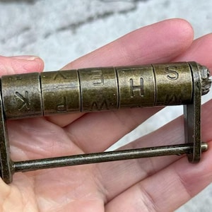 May include: Antique brass cipher wheel with rotating lettered cylinders. The letters visible are K, P, E, H, and S. The cipher wheel is held in a hand.