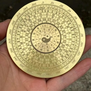 May include: A round, brass compass with intricate markings and Chinese characters. The compass features a central Yin and Yang symbol and degree markings around the perimeter. The compass is held in a hand.