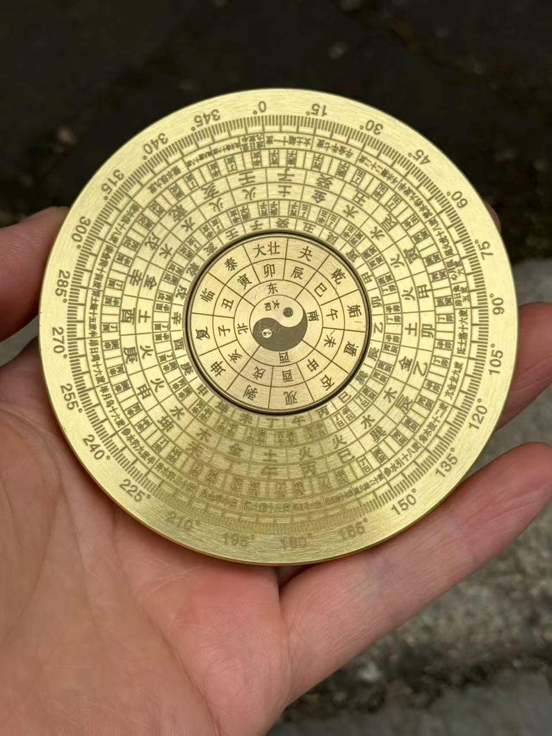 May include: A round, brass compass with intricate markings and Chinese characters. The compass features a central Yin and Yang symbol and degree markings around the perimeter. The compass is held in a hand.