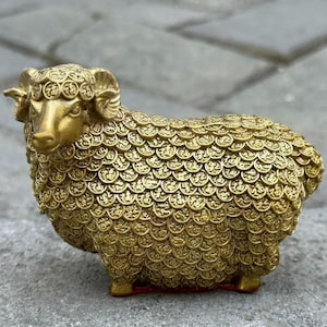 Könnte beinhalten: Eine goldfarbene Widderfigur mit aufwendigen, münzartigen Details, die ihren Körper bedecken. Der Widder hat gebogene Hörner und steht auf vier Beinen. Die Figur besteht aus einem glänzenden, metallischen Material und ist ein dekorativer Gegenstand.