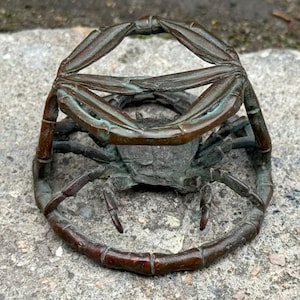 May include: A bronze-toned metal crab figurine featuring a circular frame structure. The crab is positioned in the center with its legs and claws extending outward to support the ring-shaped frame, displaying a weathered, dark patina finish.