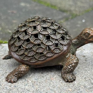 Könnte beinhalten: Eine bronzefarbene Metall-Schildkrötenfigur mit einem Panzer, der aus einem Muster überlappender kreisförmiger Münzmotive besteht. Die Schildkröte hat eine texturierte, schuppige Haut an Kopf und Beinen mit einem dunklen, patinierten Finish.