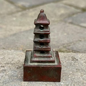 Può includere: Una piccola statuetta a forma di pagoda in metallo bruno-rossastro, caratterizzata da una struttura a quattro livelli con aperture circolari, poggiante su una solida base quadrata con una finitura patinata e usurata.
