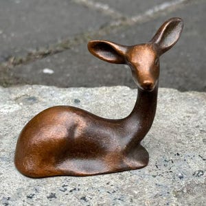 May include: A bronze deer figurine in a resting pose. The sculpture features a slender neck, large ears, and a smooth, curved body. The deer is sitting on a textured, grey surface, showcasing the detailed craftsmanship of the miniature animal.