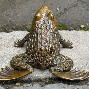 May include: A bronze frog sculpture with detailed skin texture, outstretched limbs, and webbed feet. The frog is positioned on a gray stone surface, showcasing its intricate design and metallic sheen. This decorative item is suitable for home decor.