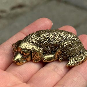 May include: A small, solid brass figurine of a dog in a curled, sleeping position. The metal surface features a polished, golden-toned finish with detailed, textured carvings that define the animal's fur, ears, and facial features in relief.