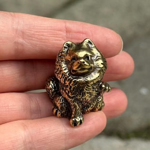 May include: A small, detailed brass figurine of a Chow Chow dog. The dog is sitting with its front paws extended, and its fur is textured. The figurine has a golden color and is likely made of metal.