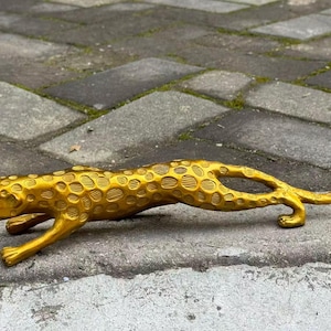 Puede incluir: Una figura de metal de color dorado que representa a un leopardo en una postura de acecho, cerca del suelo. La superficie del leopardo presenta un patrón texturizado y repetitivo de manchas circulares en el cuerpo, la cabeza y la cola.