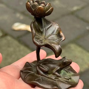 May include: A bronze frog figurine resting on a lily pad, with a lotus flower above. The sculpture is detailed with visible veins on the leaves and the frog's textured skin. The figurine is held in a hand.