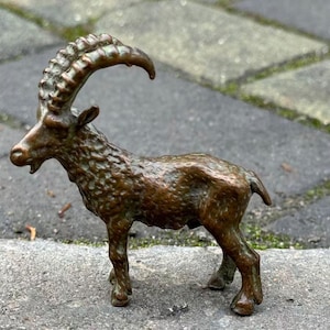 May include: A small bronze-coloured figurine of an ibex standing in profile. The sculpture features a textured coat, a short tail, and prominent, curved, ridged horns extending backwards from the head. The animal is depicted with its mouth slightly open.