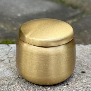 May include: A small, cylindrical container with a rounded, matching lid, crafted from a brushed gold-toned metal. The object features a smooth, uniform surface with a subtle metallic sheen and a simple, compact design with a flat base.