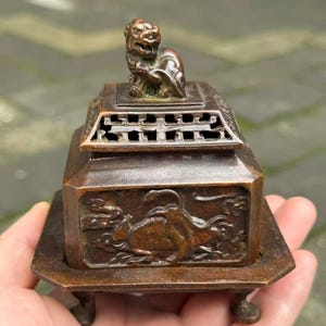 May include: A square bronze incense burner featuring a sculpted lion figure on the lid. The body displays intricate relief carvings of mythical creatures and clouds. The vessel rests on four small feet and includes a perforated lid design.