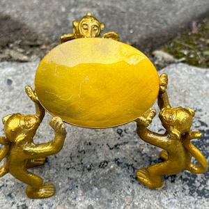 May include: A golden-colored decorative dish held by three monkey figurines. The dish is round and has a glossy finish. The monkeys are in a playful pose, supporting the dish with their arms. The overall theme is decorative.