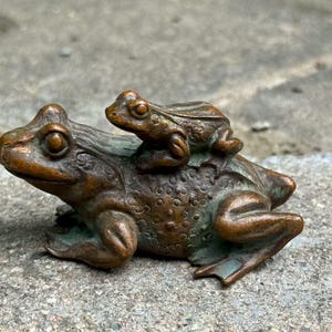 Puede incluir: Escultura de rana de bronce que representa una rana más grande con una rana más pequeña sobre su espalda. La escultura tiene una textura detallada y un aspecto envejecido, con toques de pátina verde. Las ranas están representadas en un estilo naturalista.