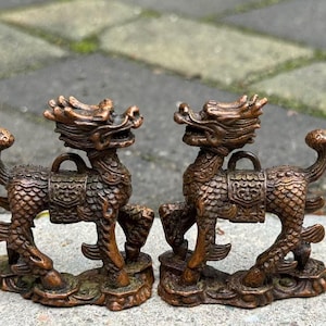 Może przedstawiać: Dwie figurki mitycznych stworzeń w kolorze brązu. Każda figurka ma smoczą głowę, siodło i ogon zakrzywiony do góry. Figurki stoją na podstawie o szczegółowym, teksturowanym wzorze. Ogólna estetyka jest antyczna i szczegółowa.