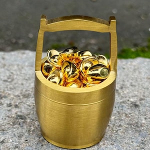 Peut inclure: Un petit seau doré avec une poignée, rempli de nombreux objets dorés en forme de capsule. Le seau a un corps cylindrique et une poignée incurvée. Les objets à l'intérieur semblent réfléchissants, captant la lumière.