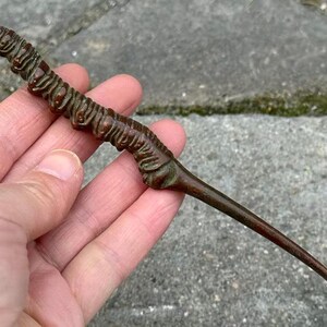 May include: A bronze-colored, worm-shaped sculpture with a long, curved tail. The sculpture has a textured surface with detailed ridges and a slightly green hue in some areas. The object is held in a hand.