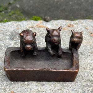 Può includere: Una scultura in bronzo raffigurante tre piccoli maiali in una mangiatoia. I maiali sono rivolti in avanti, con dettagli precisi. La mangiatoia è rettangolare e ha un aspetto scuro e invecchiato. La scultura è su uno sfondo sfocato.