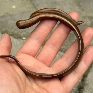 Può includere: Un bracciale aperto e curvo realizzato in un materiale marrone scuro con una finitura liscia e lucida. Il design presenta un'estremità affusolata e appuntita e una sezione arrotondata più spessa, creando una forma fluida e organica.