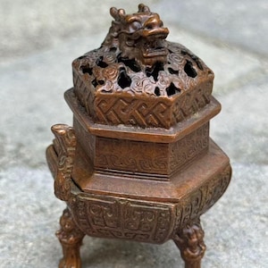 May include: A bronze-colored metal incense burner with a hexagonal body and three legs. The lid features an openwork pattern and a sculpted dragon head at the top. The sides of the burner are decorated with intricate geometric and relief carvings.