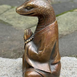 Puede incluir: Una escultura de tono bronce de una figura reptiliana con cabeza de serpiente y escamas texturizadas, que viste una túnica. La figura está sentada con las manos juntas en un gesto de oración, sosteniendo un pequeño collar de cuentas.