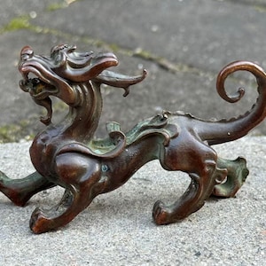 Może przedstawiać: Brązowa figurka mitycznego stworzenia, być może smoka. Rzeźba ma szczegółowy wzór ze zwiniętym ogonem i otwartą paszczą. Kolor to mieszanka brązu i zieleni.