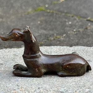 May include: A bronze-colored sculpture of a dog in a resting pose. The dog is lying down with its front paws extended and its head raised. The sculpture has a smooth, polished finish and is set against a gray, textured surface.