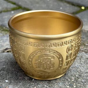 May include: A metallic gold-colored bowl with a smooth interior and a textured exterior featuring embossed Chinese characters and a geometric border pattern. The bowl has a rounded shape and a matte finish, displaying intricate relief designs.
