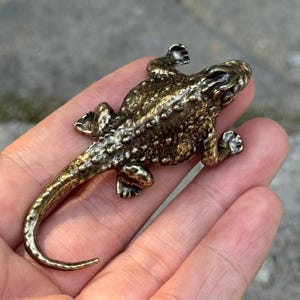 May include: A small, metallic bronze-colored lizard figurine with a textured, bumpy surface. The lizard is depicted in a crawling position with its long, curved tail extending downward. The finish has dark highlights in the crevices.