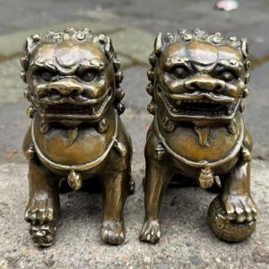 May include: Two bronze-colored Foo Dog statues. Each statue has a detailed, fierce expression, with intricate mane and facial features. They are posed in a seated position, with one paw resting on a ball. The statues are likely used for decorative purposes.