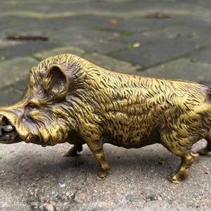 May include: A metallic gold-colored figurine of a wild boar standing in profile. The sculpture features detailed, textured fur, a prominent snout with visible tusks, pointed ears, and a small, curled tail. The surface has a polished, reflective finish.